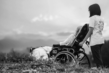 Ältere Person von hinten im Rollstuhl betreut von junger Pflegerin mit dunklen langen Haaren in der Natur vor einer Bergkulisse.